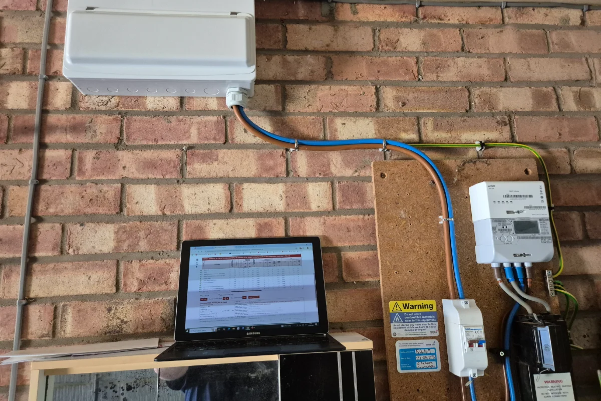 Electrical metering equipment mounted on a brick wall beside a laptop displaying system data.